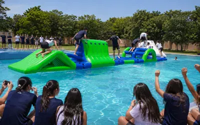 Alumnos de secundaria cruzando una pista de obstáculos inflable dentro de la alberca de su escuela, con sus compañeros animando desde la orilla.