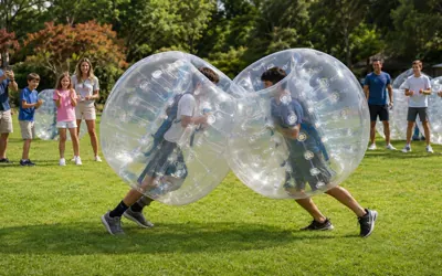 Dos jóvenes chocando dentro de bumper balls inflables transparentes sobre el pasto, mientras su familia los anima en un parque.